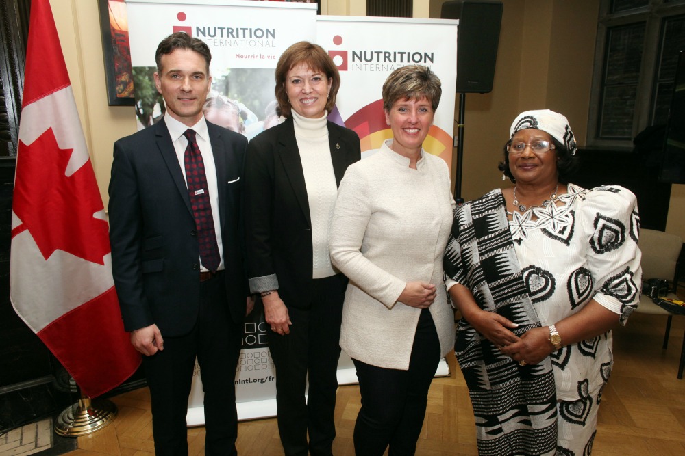 Four people standing inside, beside Canadian flag - from left to right: Joel Spicer, President and CEO of Nutrition International; Pam Damoff, Liberal Member of Parliament; the Honourable Marie-Claude Bibeau, Minister of International Development and La Francophonie; Her Excellency Dr. Joyce Banda, former President of Malawi.
