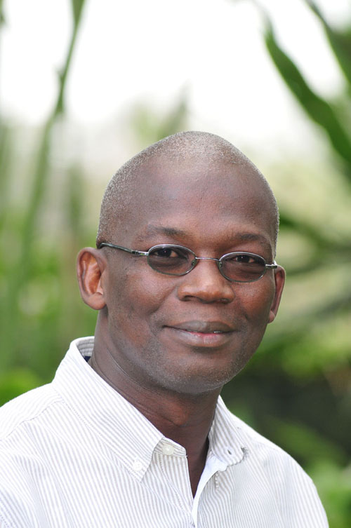 A man with glasses smiling and looking at the camera with a blurred green background behind him.