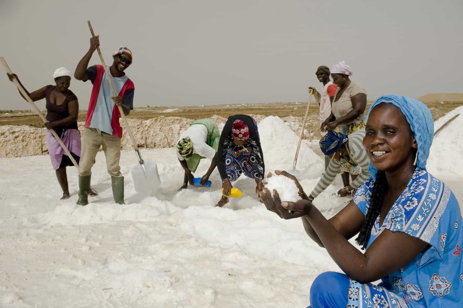 woman holding pure salt in her hand while standing in a salt pond with salt harvesters working behind her