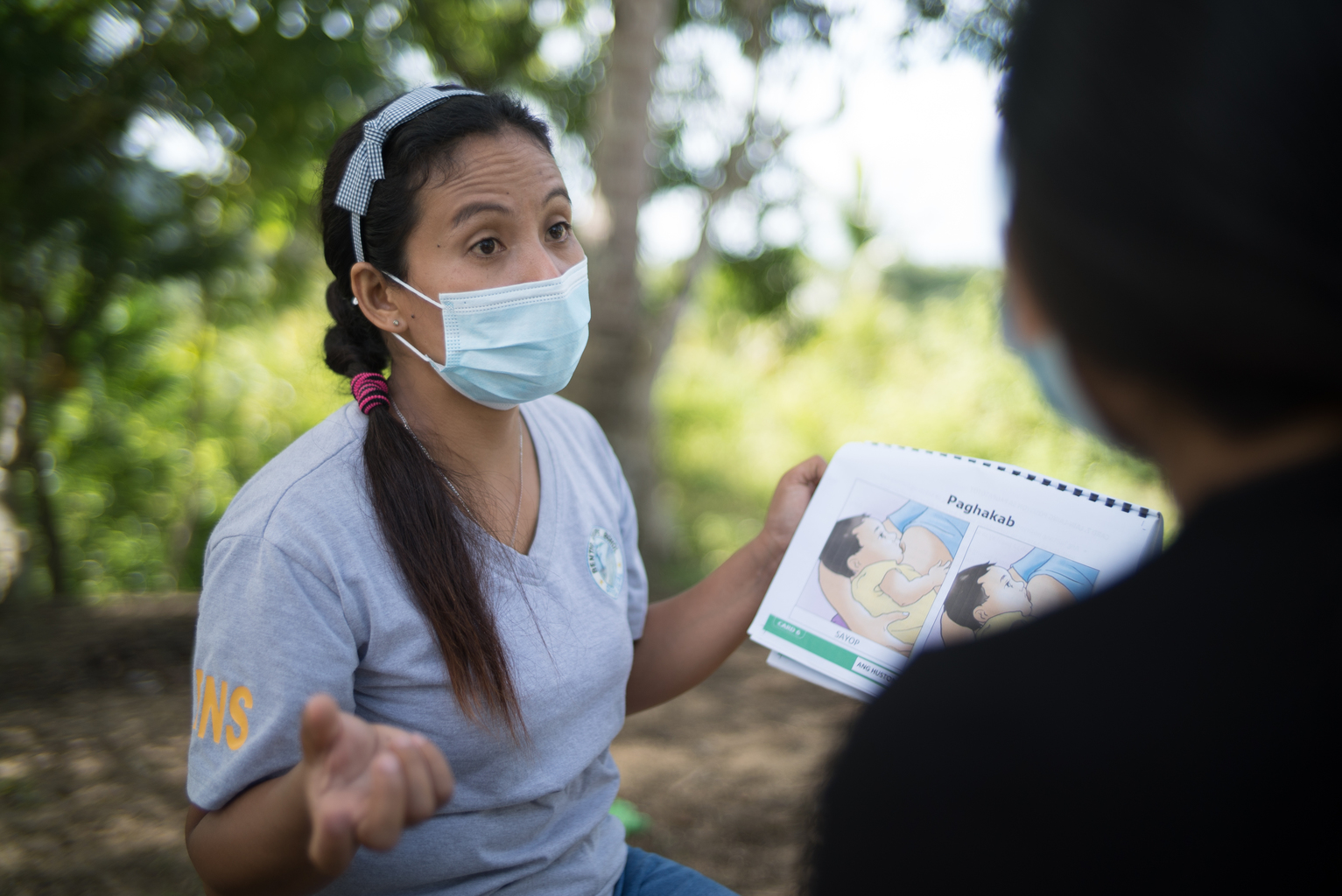 Woman wears a mask outside and holds a booklet during a mother-to-mother support group session in the Philippines