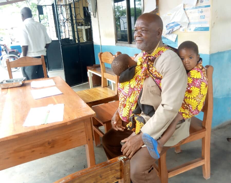 Father sits on a chair at a health clinic carrying his son on his front and holding his daughter behind him.