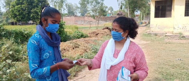 A woman standing on the right in a pink kurta and wearing a mask hands a package of iron and folic acid supplements to an adolescent girl standing on the left who is also wearing a mask.