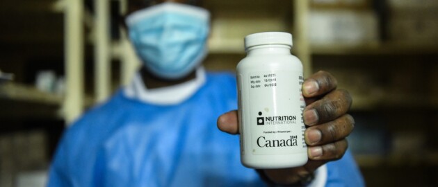 A health care working wearing scrubs and a mask holds up a bottle of vitamin A tablets