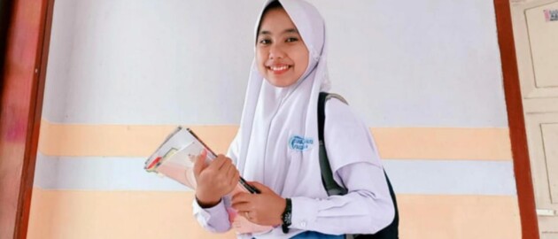 Student in Indonesia stands holding books in her hand and wearing her backpack while smiling to camera.