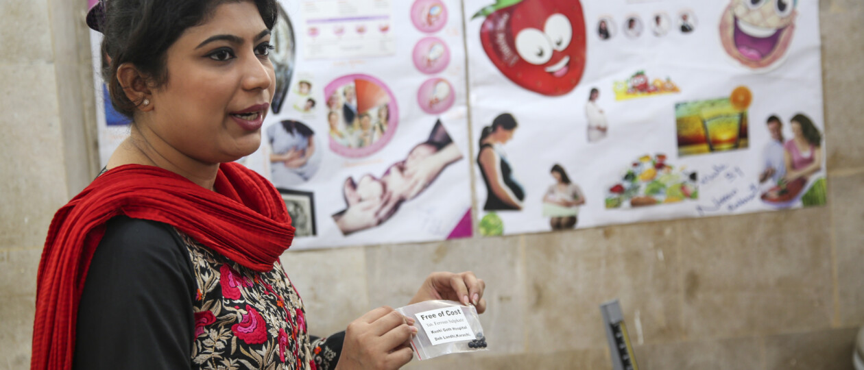 female health worker in Pakistan discussing maternal supplementation before an informational poster on antenatal care