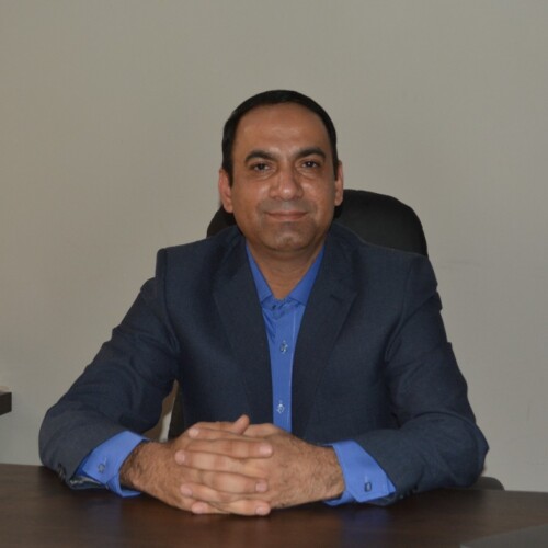 A man in a suit sitting at a desk looking directly a the camera with his hands clasped.