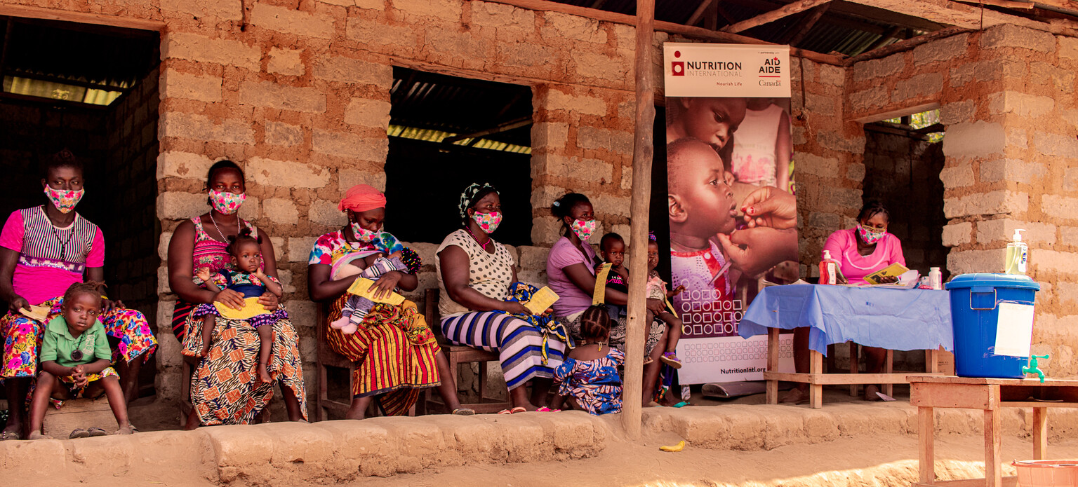 Mothers wait outside a health post on wooden chairs with their children in their laps as the nurse completes paperwork to prepare their children for a vitamin A dose.