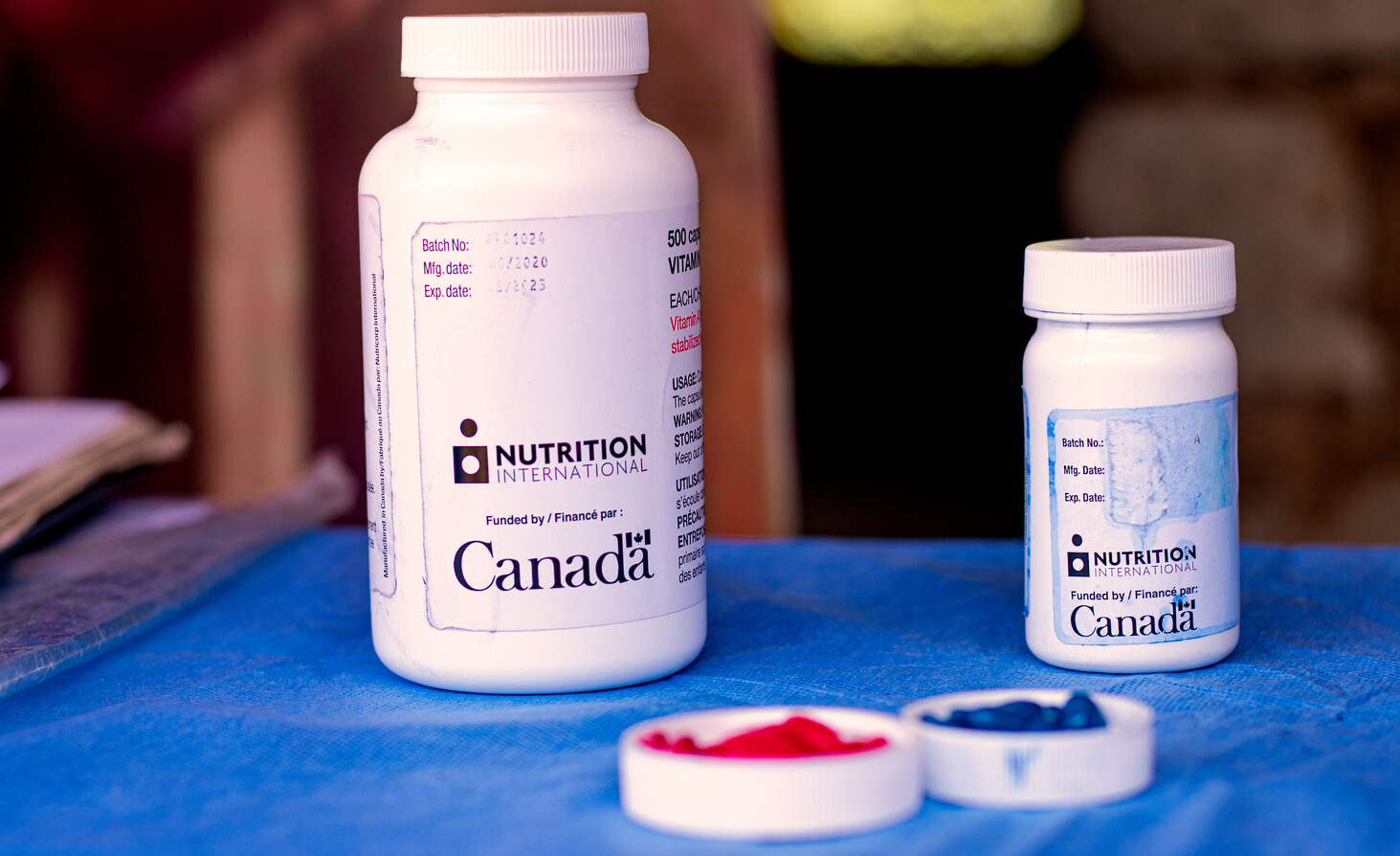 Blue and red vitamin A capsules lie in a bottle lid on top of a blue table alongside the Nutrition International and Government of Canada branded bottles.