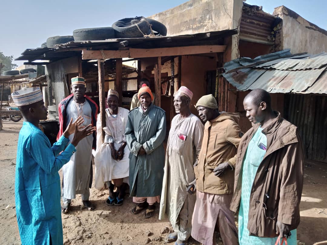 A man stands with his back to the camera speaking to a group of men outside who are listening intently in Nigeria