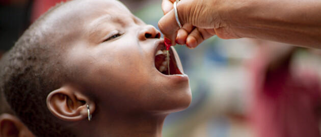 School aged girl in the DRC hold her mouth open while a red capsule of vitamin A supplemenration is administered orally.