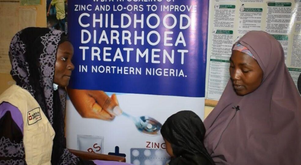 Thirty-four-year-old mother sits inside a a health centre with her fifteen-month-old-baby on her lap as a health worker sits beside her and counsells her on diarrhoea management and zinc and LO-ORS treatment.