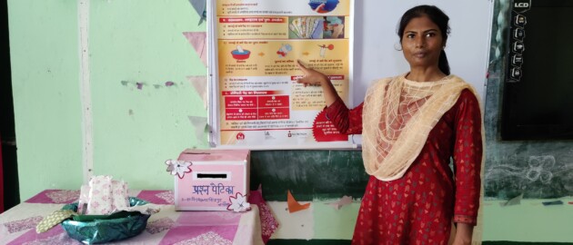 Teacher in a red kurta stands in front of a poster in a classroom. She is pointing at the poster as she looks at the camera.