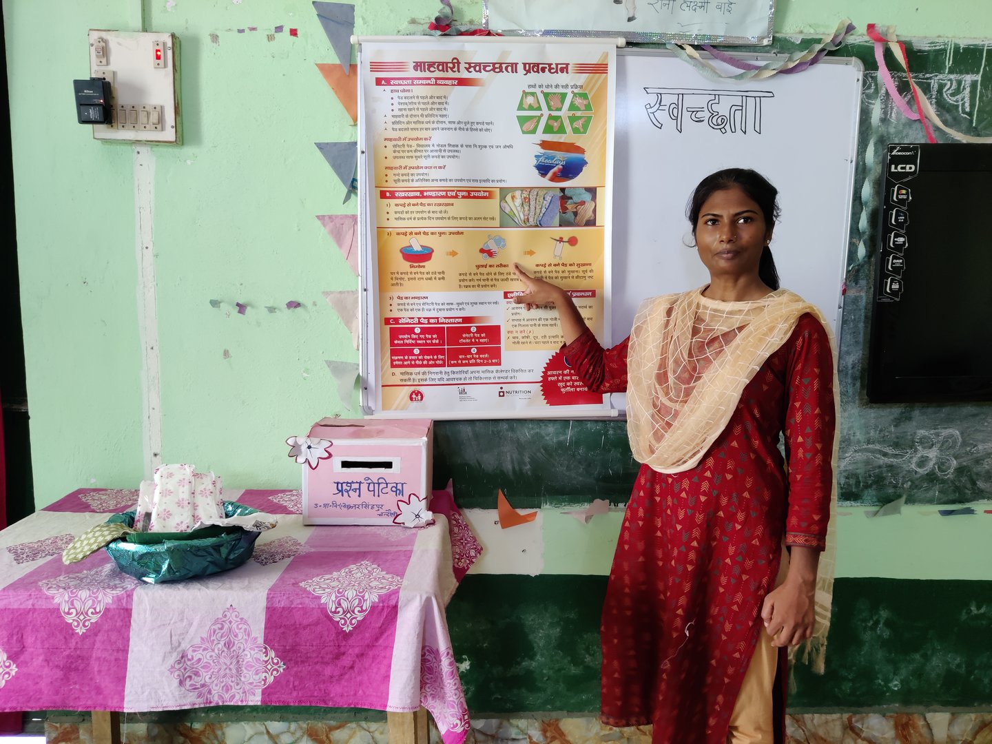 Teacher in a red kurta stands in front of a poster in a classroom. She is pointing at the poster as she looks at the camera.