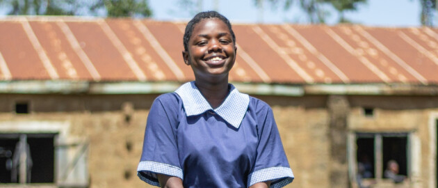 A female student wears a blue school uniform and stands outside in a grass field.