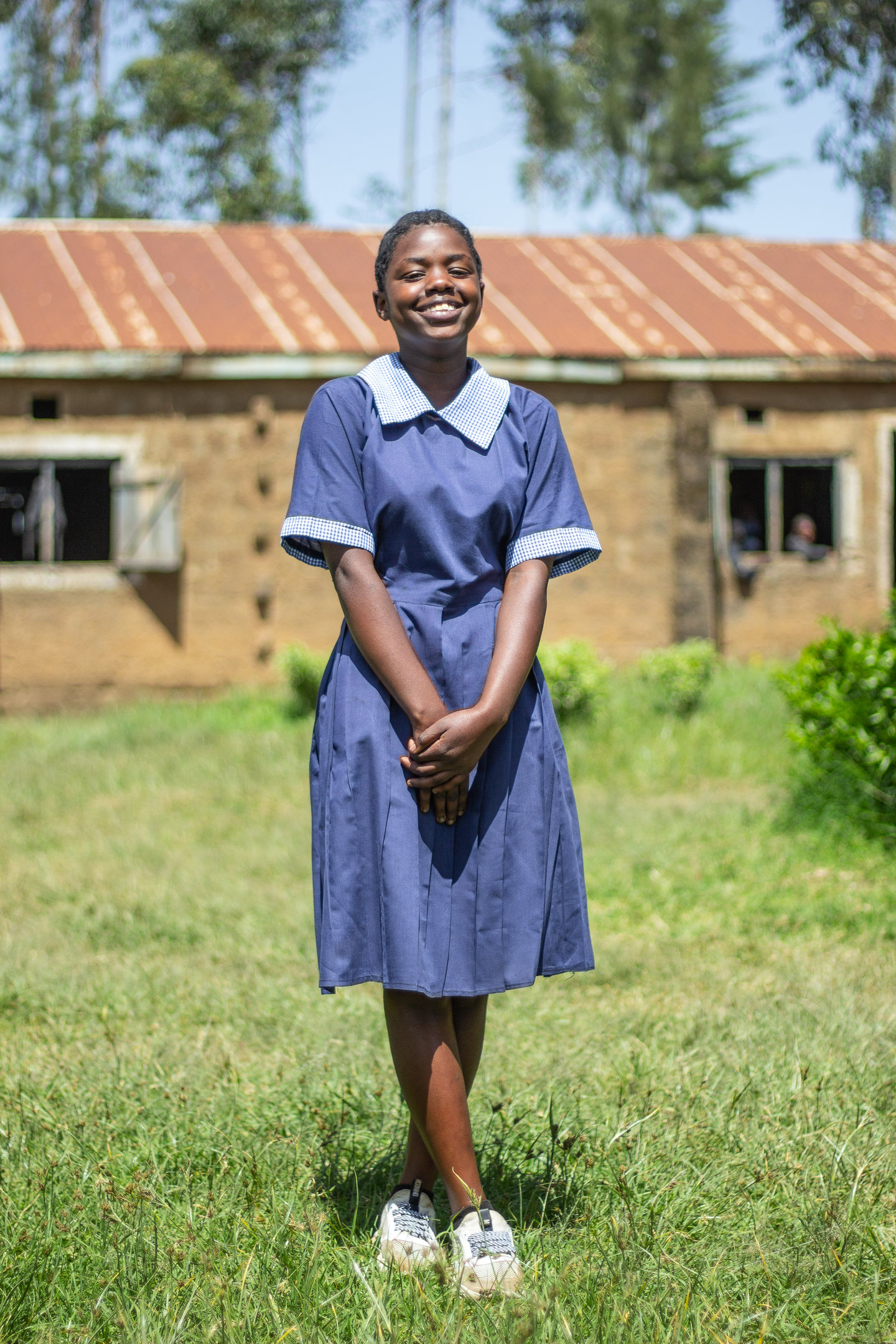 A female student wears a blue school uniform and stands outside in a grass field.