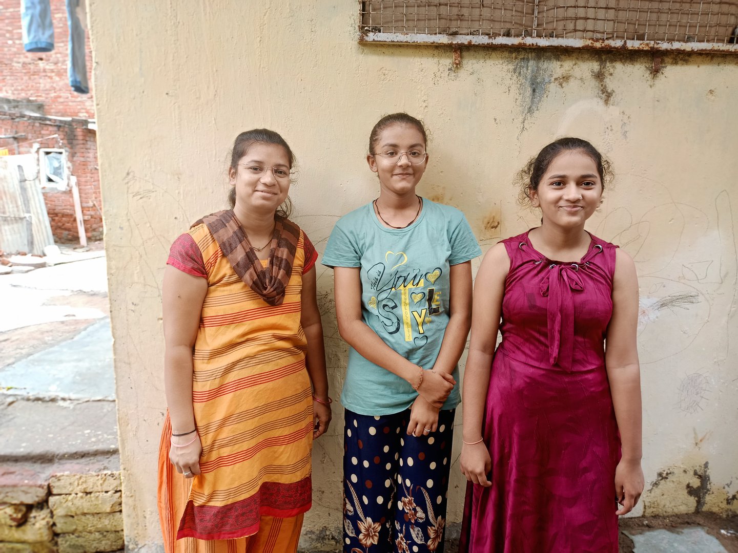 Three people stand in of a wall smiling at the camera in India.