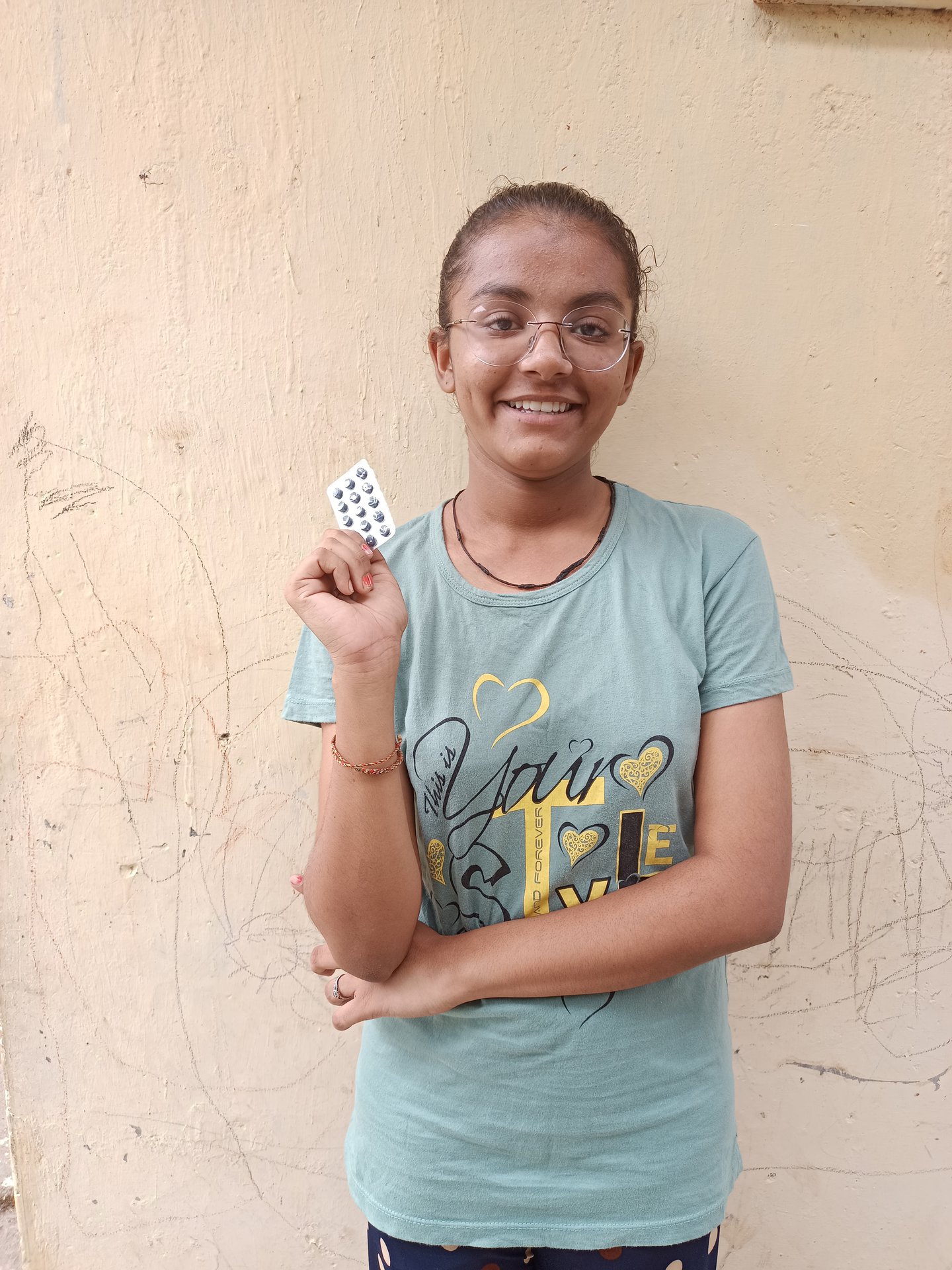 An adolescent girl stands infront of wall holding a package of weekly iron and folic acid supplementation to prevent anaemia. She is smiling to camera.