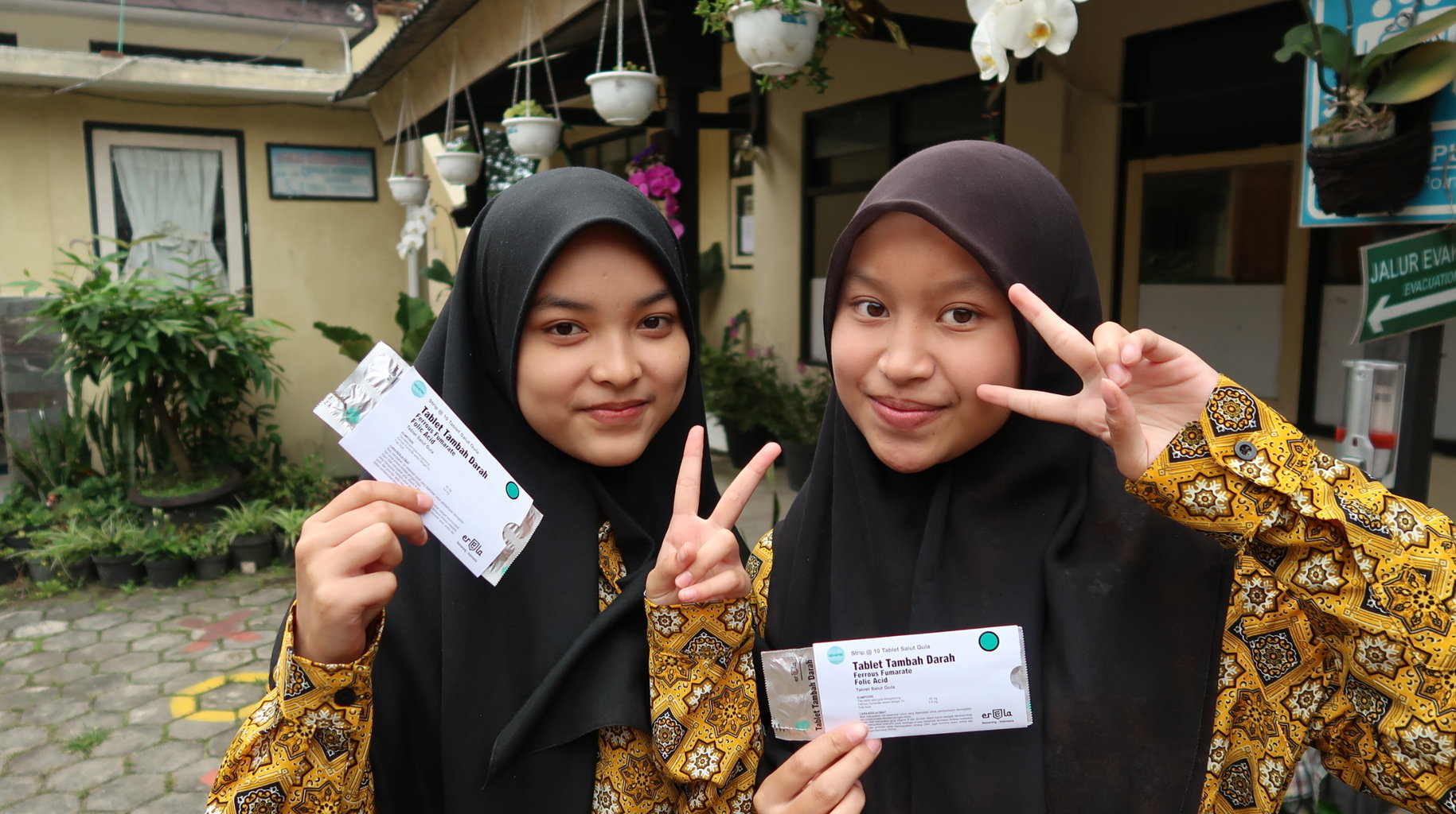 Two girls smile at the camera giving peace signs and holding up packages of weekly iron and folic acid supplementation.