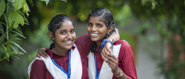 Two girls in school uniforms laugh to the camera with their arm around their friend.