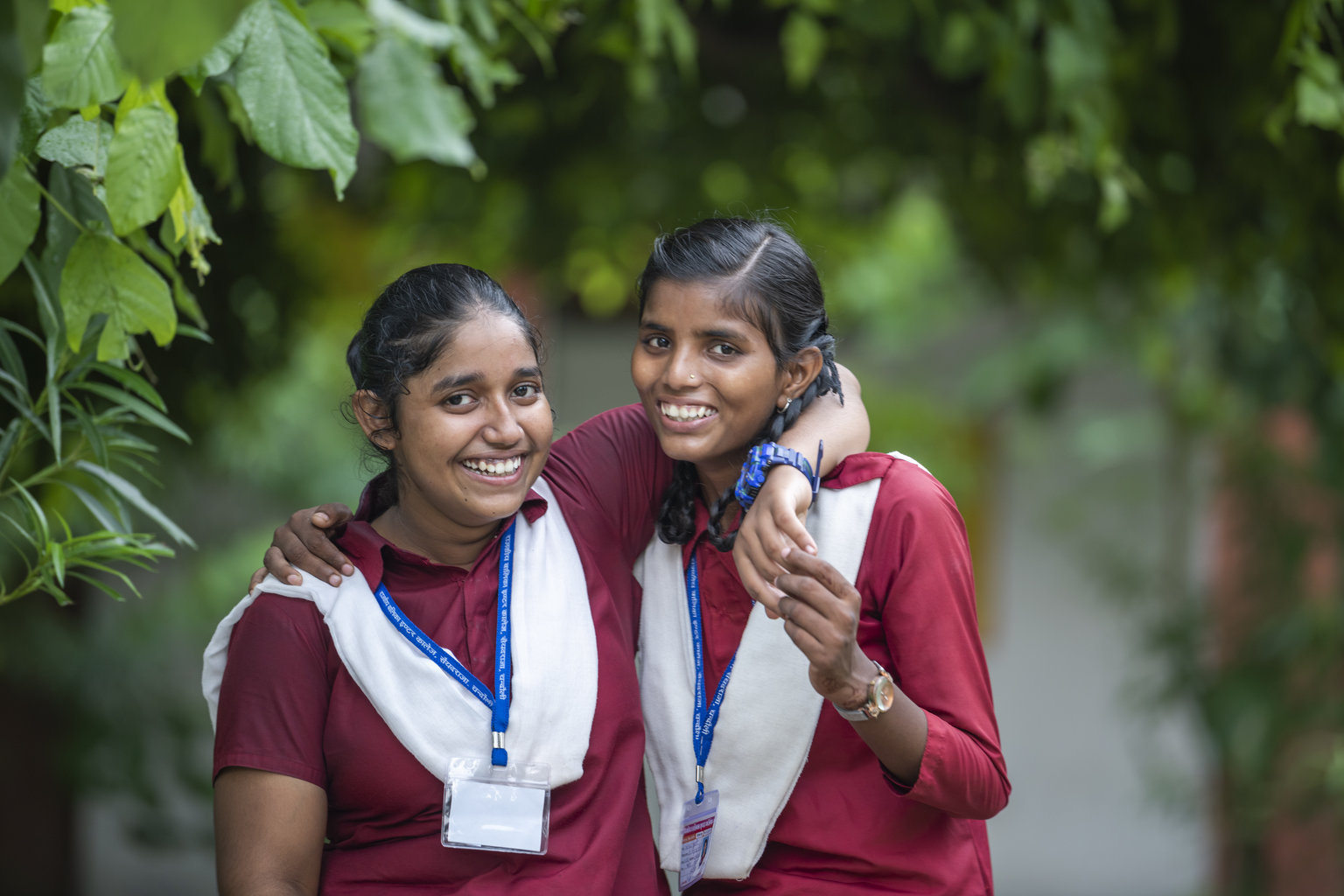Two girls in school uniforms laugh to the camera with their arm around their friend.