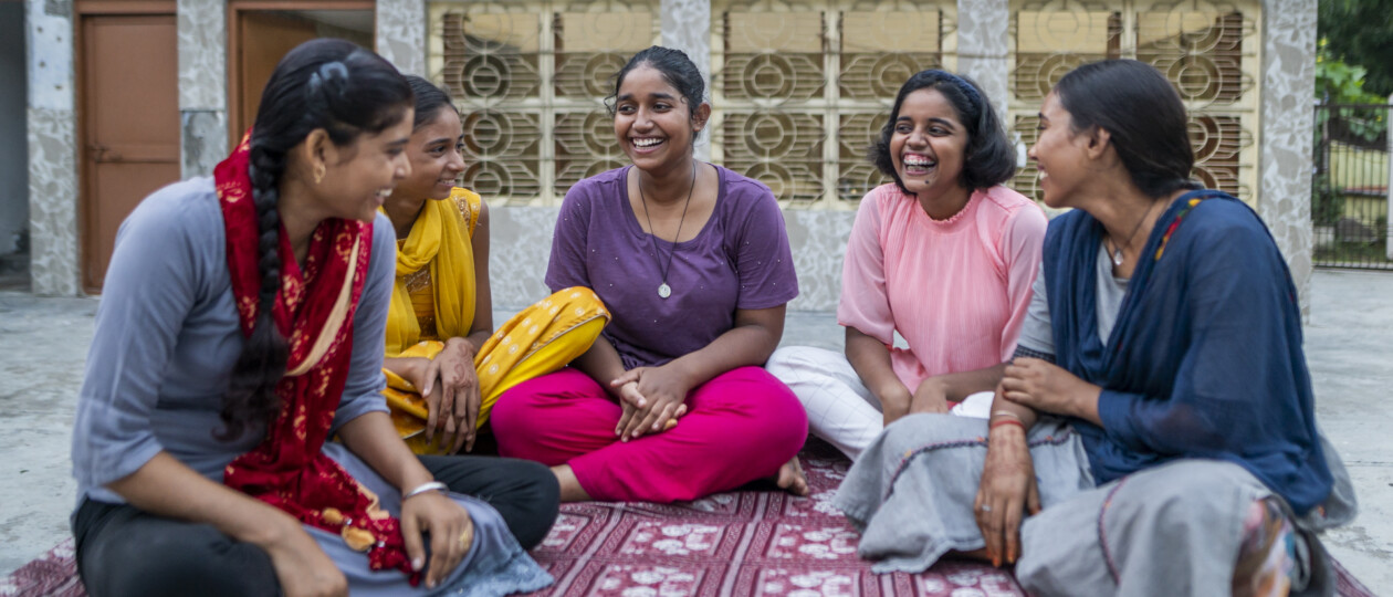 Girls sit in a circle on a blanket smiling and laughing together.
