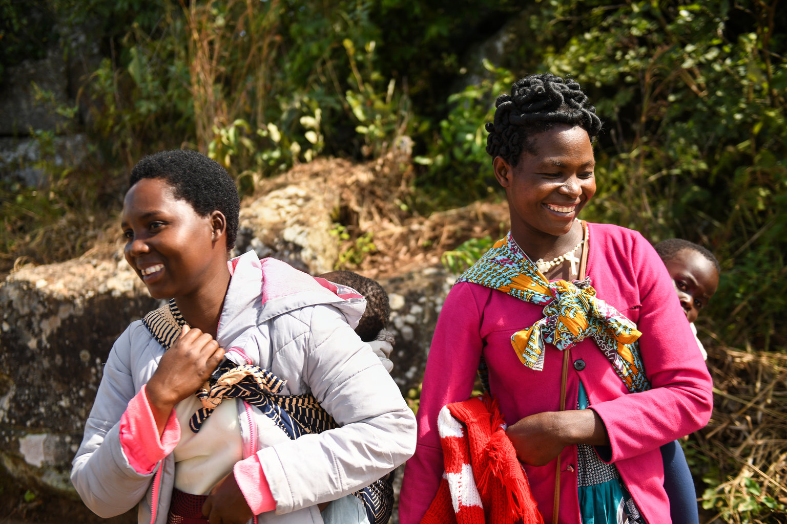 Two women laugh with babies on their backs wearing bright clothing.