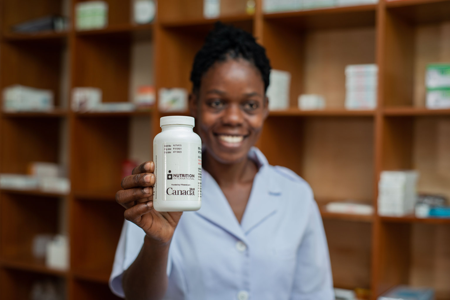 A woman is standing in a pharmacy holding up a vitamin A supplementation bottle smiling to the camera.