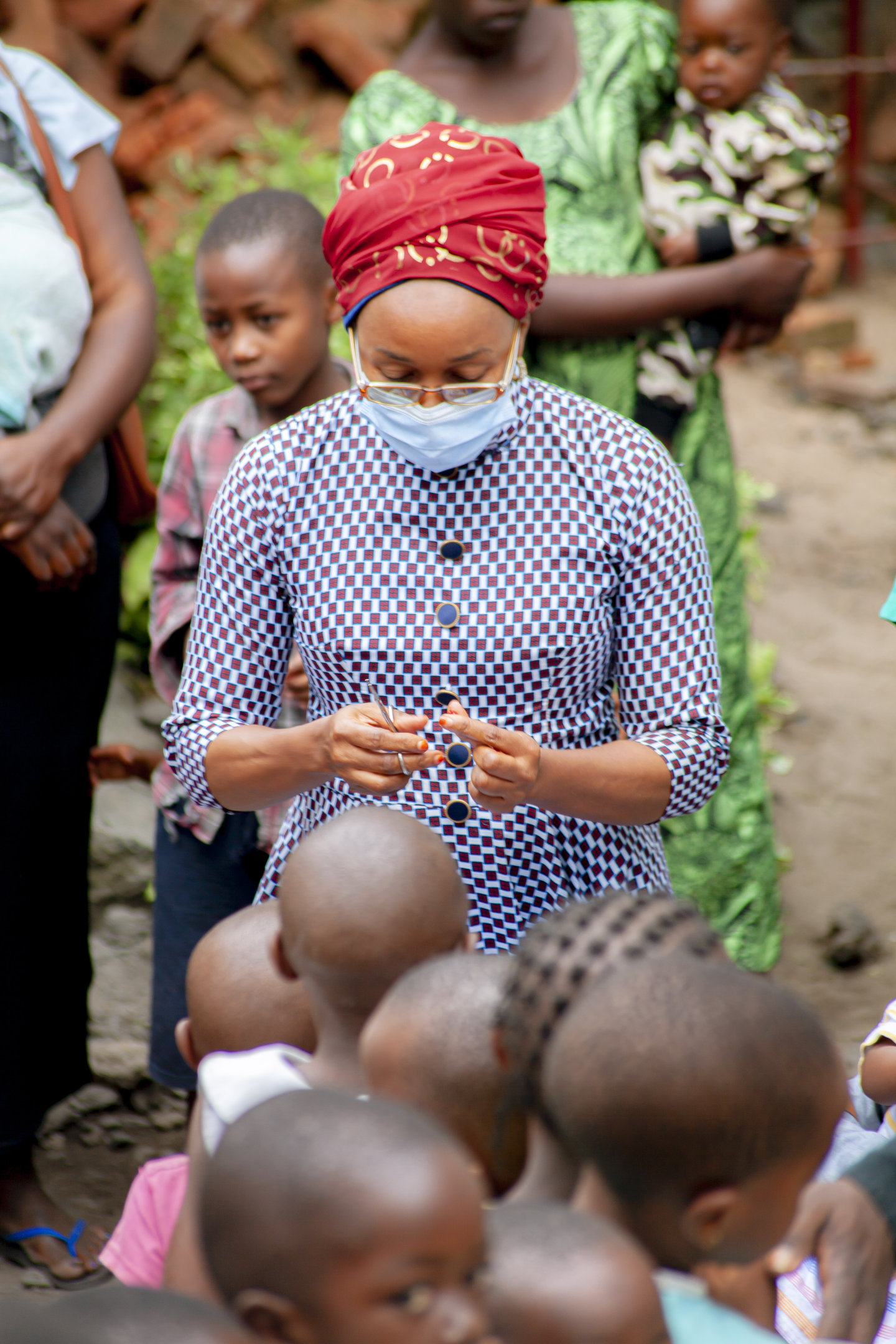A woman in a checkered blue dress wearing a medical mask cuts open a vitamin A capsule to give to young children who are lined up in front of her.