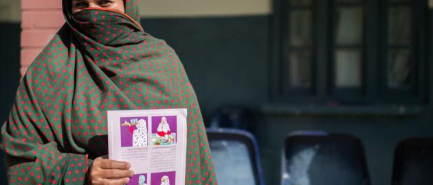 A woman stands outside wearing a niqab that is green and red. She is holding a maternal nutrition pamphlet that has illustration on when and how to consume multiple micronutrient supplements.