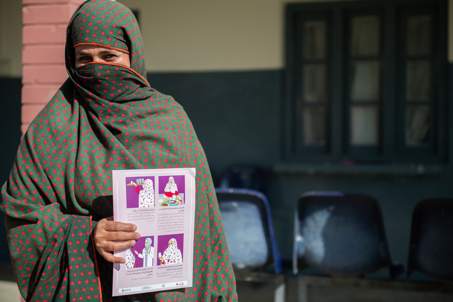 A woman stands outside wearing a niqab that is green and red. She is holding a maternal nutrition pamphlet that has illustration on when and how to consume multiple micronutrient supplements.