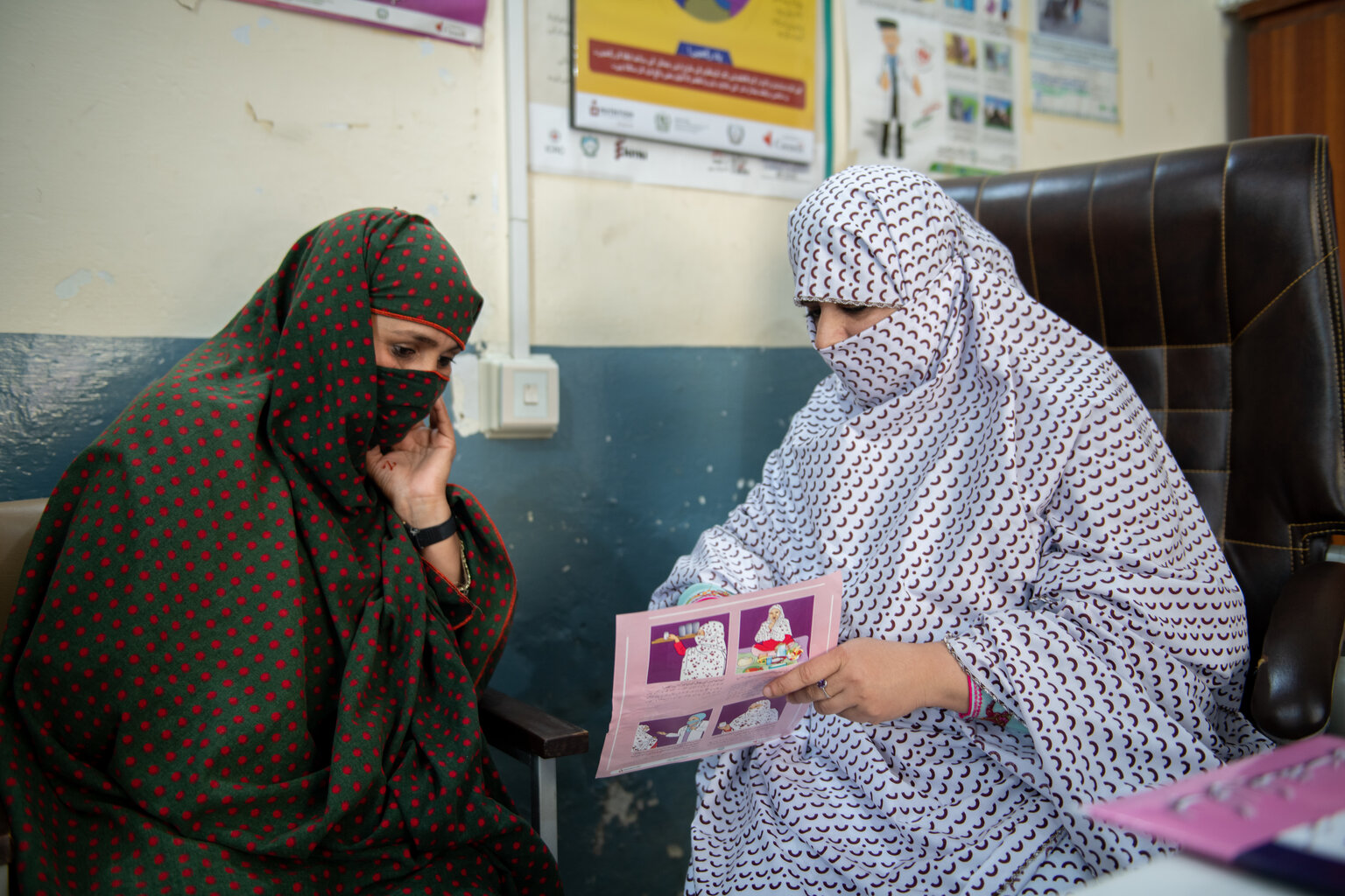 Two women sit beside each one. The woman on the left is wearing a green and red niqab and listening intently to the woman on the right who is wearing a white and red niqab. She is speaking and gesturing to a pamphlet on maternal nutrition.