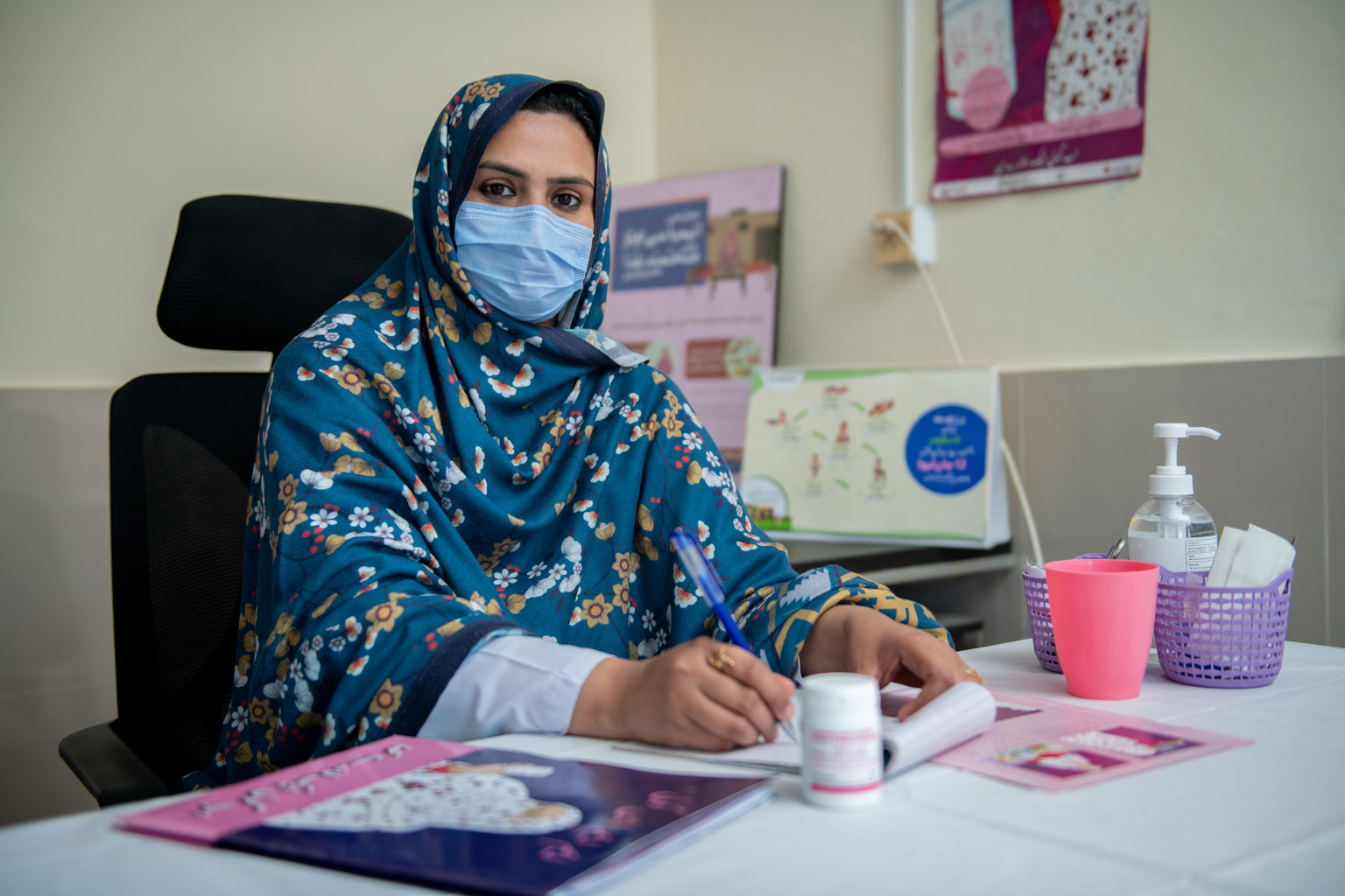 A woman sits behind a desk wearing a mask and looking directly to the camera. She is holding a pen and has information on maternal nutrition on her desk.