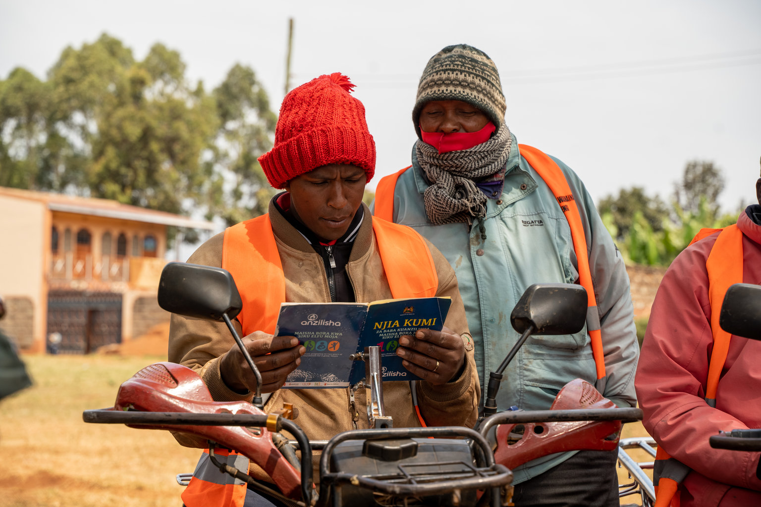 Un homme coiffé d’un bonnet rouge lit une brochure éducative tandis qu’un autre homme lit par-dessus son épaule.
