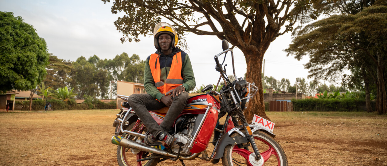 A man in an orange vest wears his helmet sitting on top of a motorcycle that he drives as a taxi service.