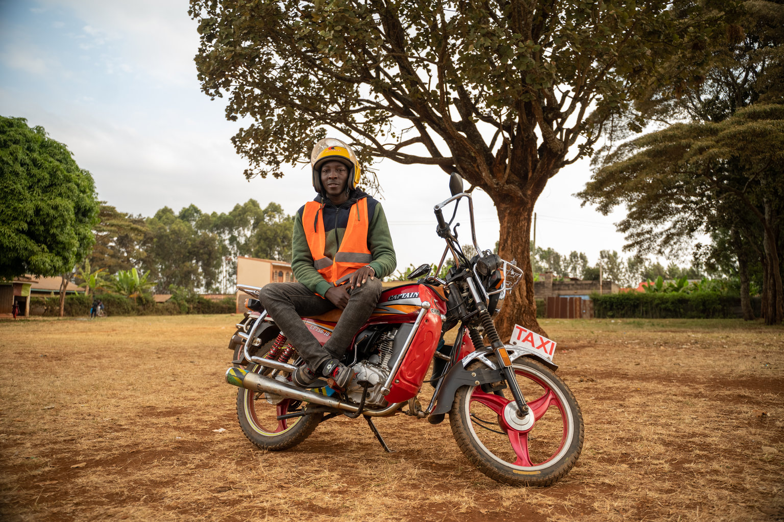 A man in an orange vest wears his helmet sitting on top of a motorcycle that he drives as a taxi service.