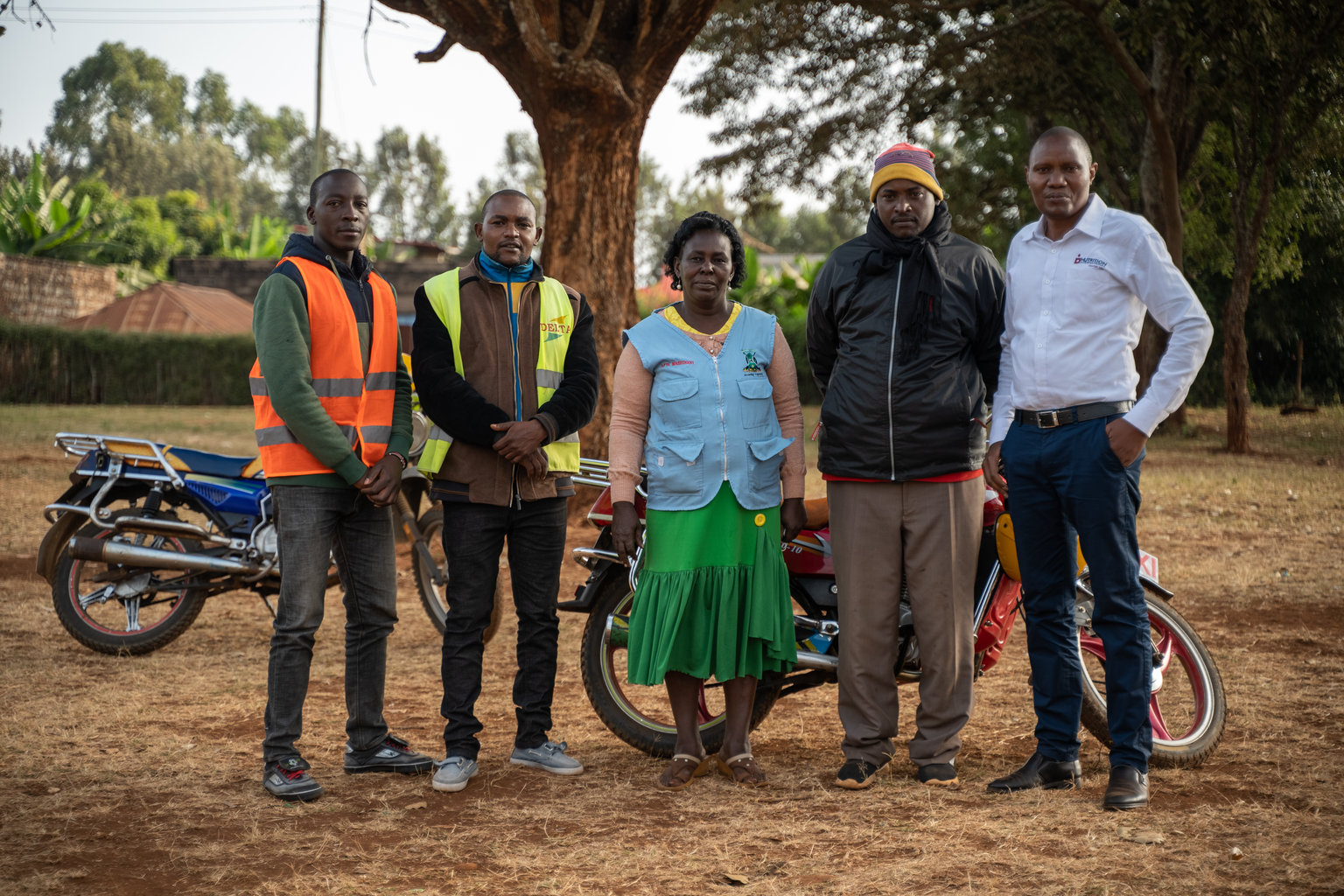 Five adults stand in a row in a field looking at the camera. A couple motorcycles are in the background.