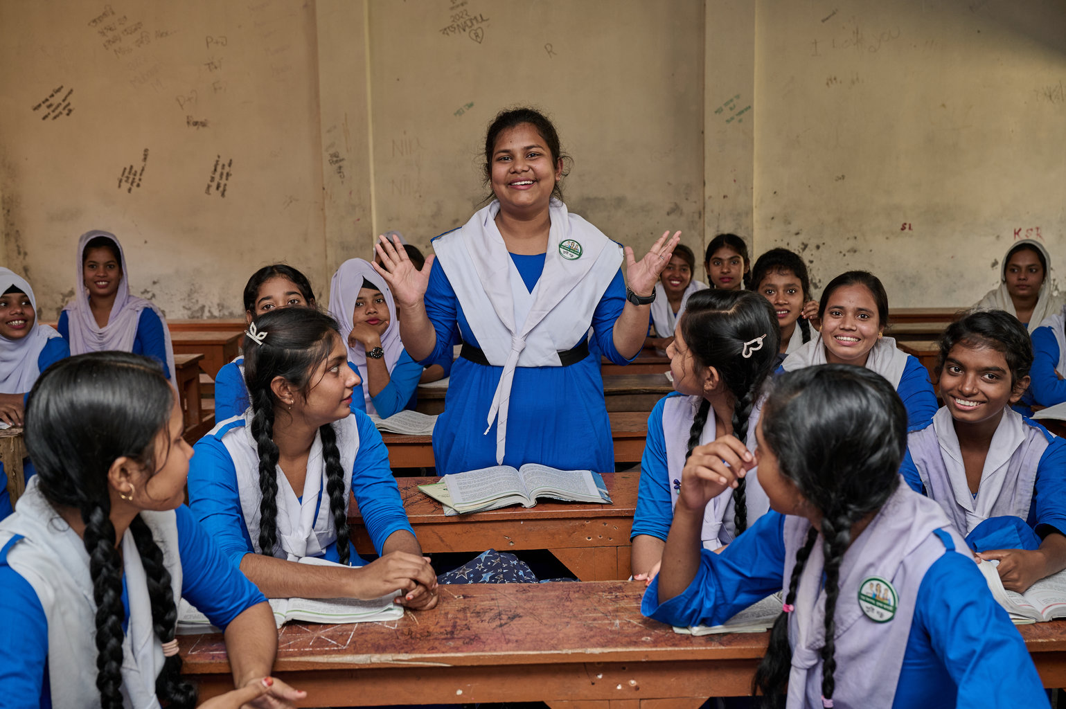 A girl stands up in the middle of the classroom. Her classmates are all turned and looking at her as she smiles and has her hands raised during a talk.