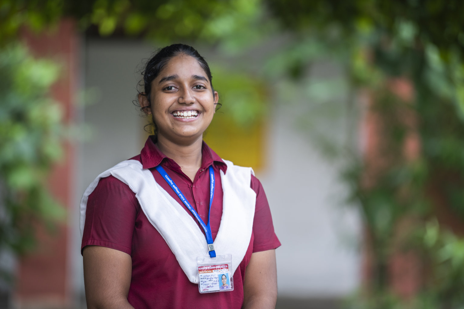  A teenage girl smiles to the camera. She is wearing her school uniform and is standing outside her school.