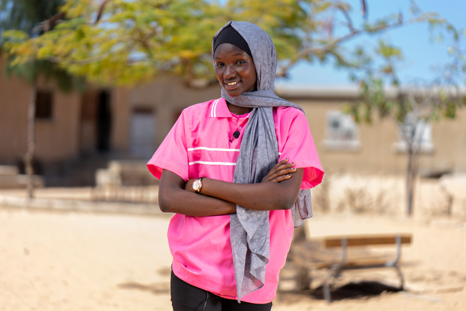 A teenage girls smile to the camera. She has her arms folder in front of her and is standing outside her school.