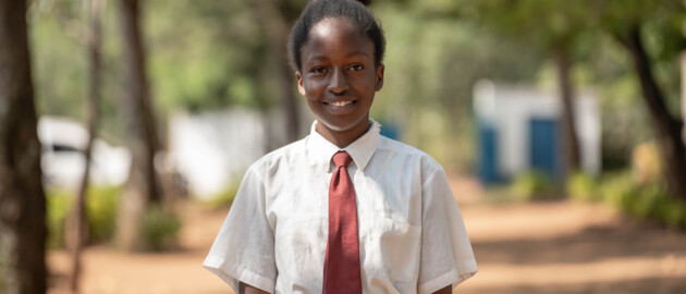 An adolescent girl in a school uniform stands outside with trees in the background. She is smiling to camera and standing directly in the middle of the frame.