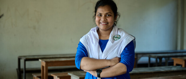 A teenage girl standing in a classroom in her school uniform. her arms are crossed in front of her and she's smiling to the camera.
