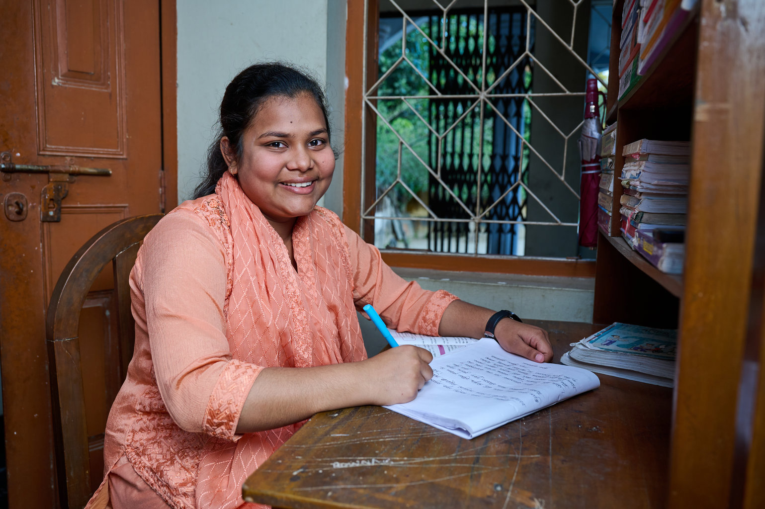 A teenage girl smiles to camera. She is holding a pen and looking up from writing in her notebook.