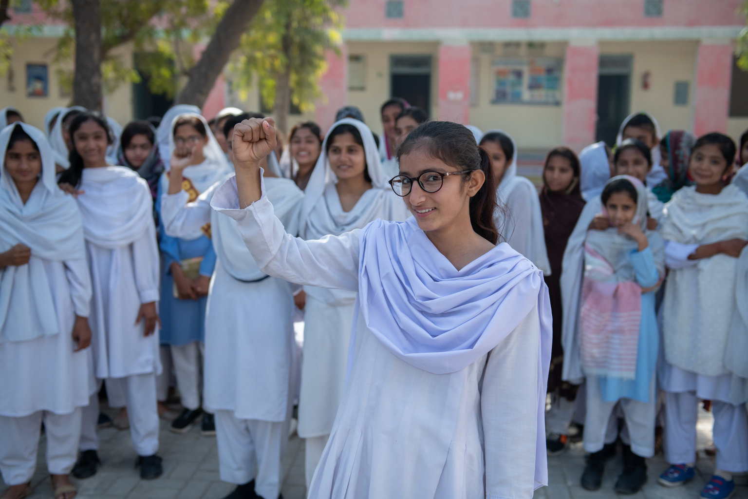 A girl raises her hand up the sky. She is standing in front of a large group of girls who are also facing the camera. They are all standing in front of their school.