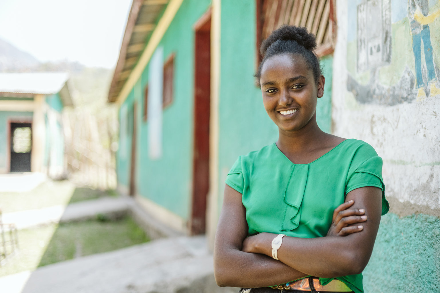 A teenage girl stands outside her classroom. She is smiling to camera with her arms crossed in front of her.