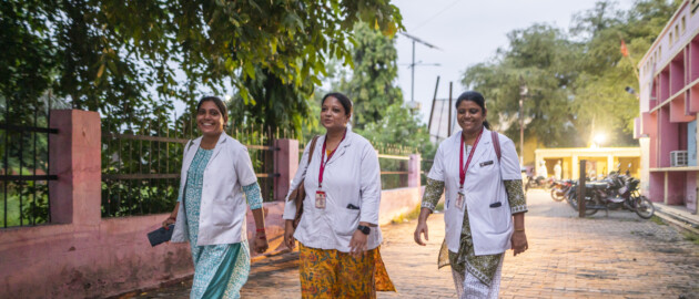 Three women wearing medical coats walk towards a hospital in India