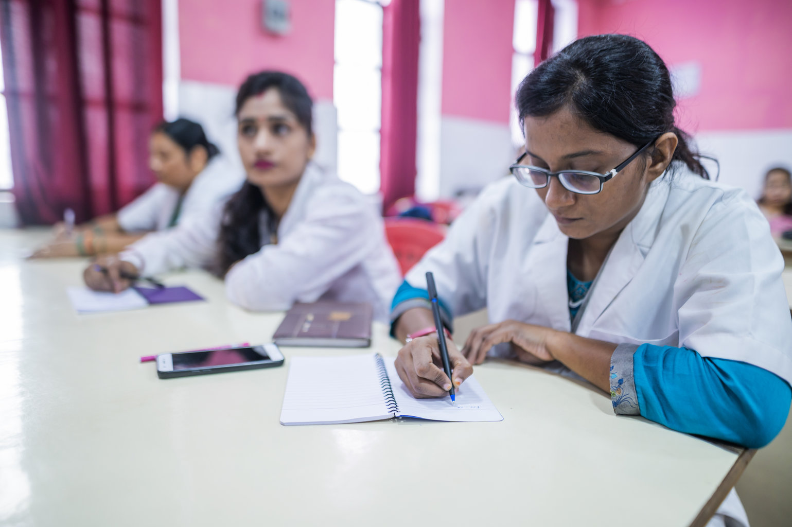 Women wearing medical coats take notes while listening to a presentation at a hospital