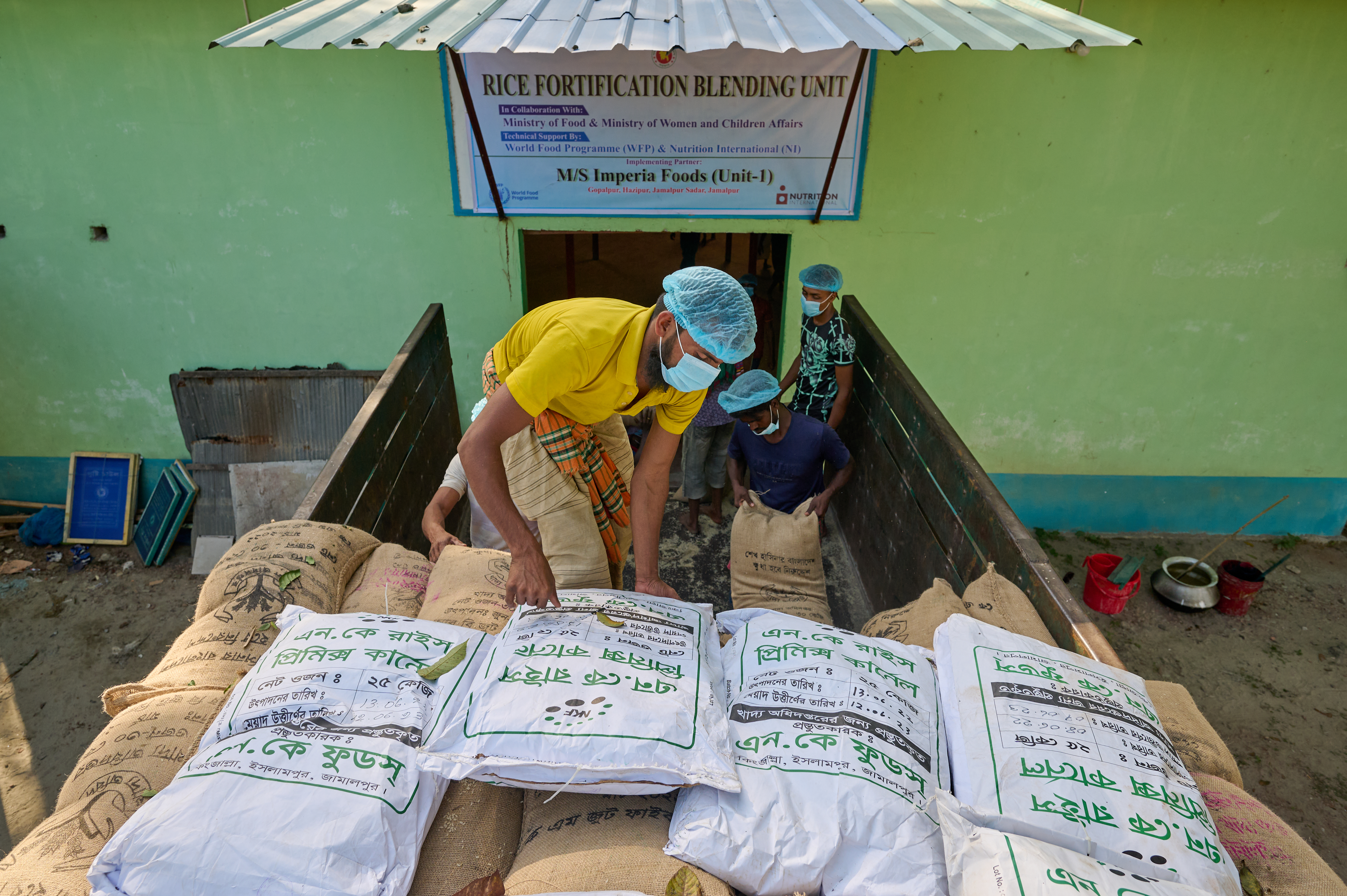 A rice fortification blending facility combines fortified rice kernels with non-fortified rice in the regulated ratio. Here, the rice is arriving on the back of a truck to be blended. Workers are unloaded the sacks of rice and carrying them into then blending facility. 
