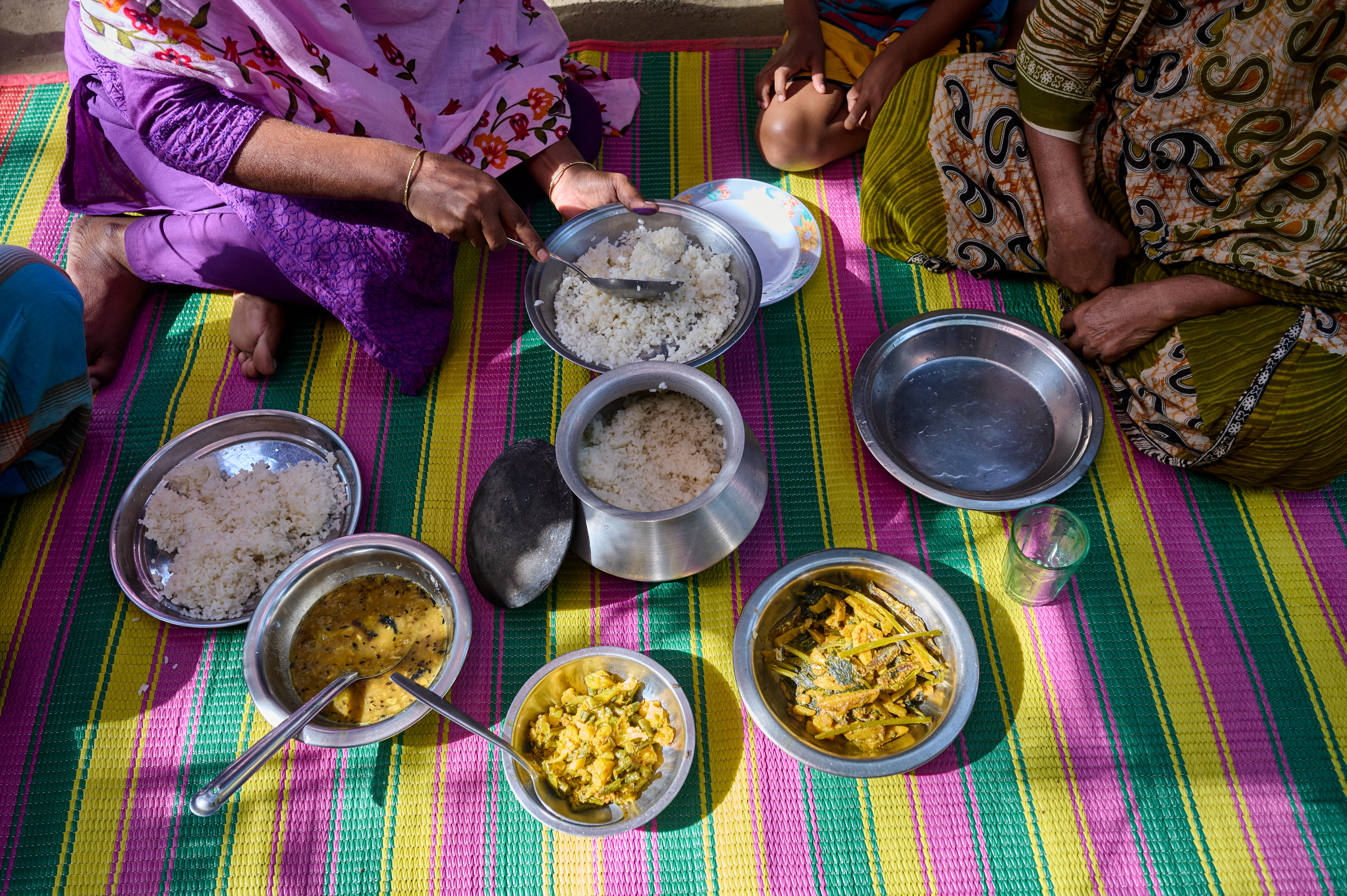 A midday meal is laid out on a colourful mat. Bowls of rice, and differnt kinds of vegetable curry are present. Two women sit cross legged on the floor, as one woman starts to serve the rice.