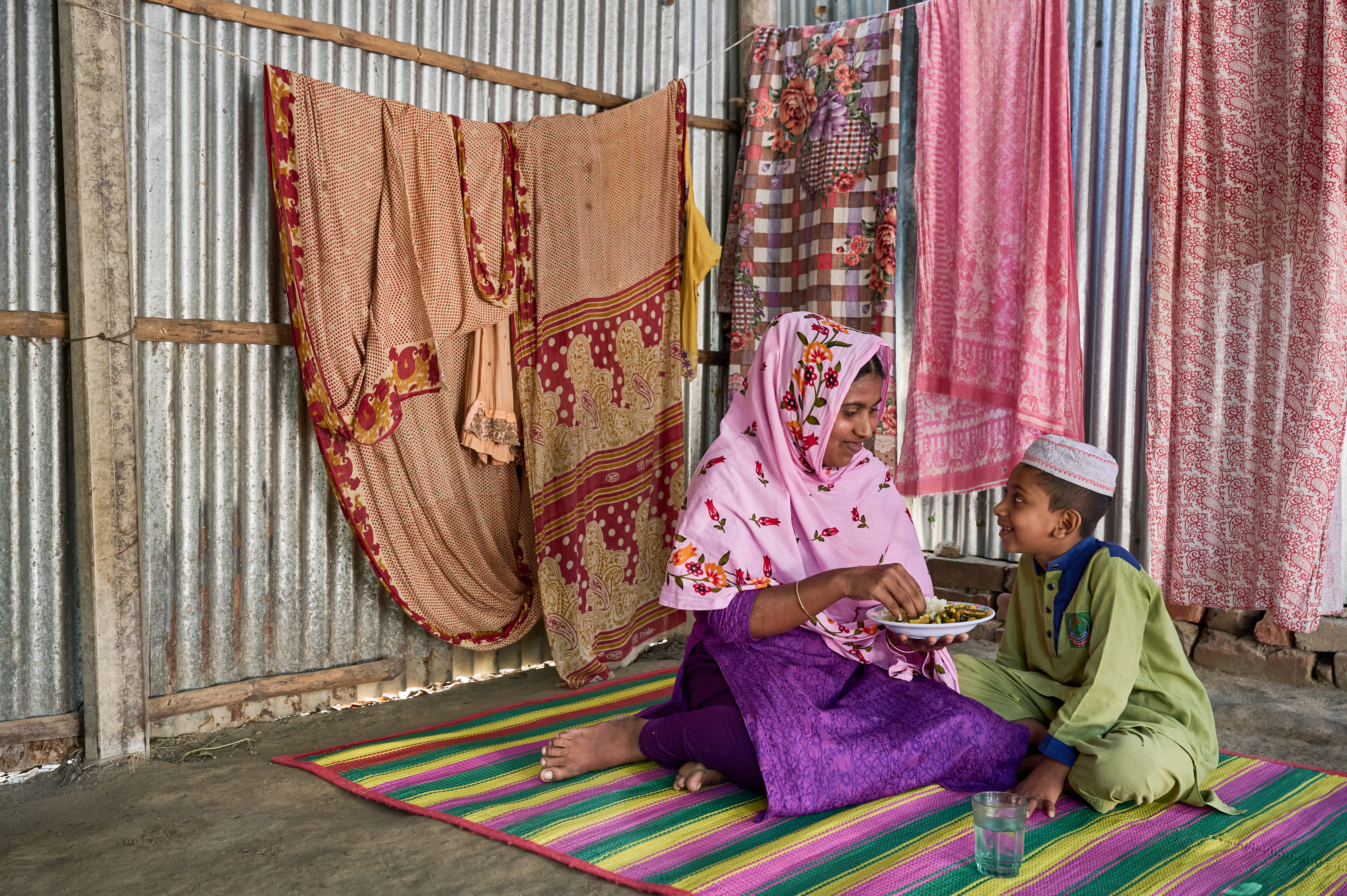 A mother and her son sit on a colourful mat in their home. The mother is feeding her son, a child under 10 years old, a lunch that includes rice fortified with micronutrients.