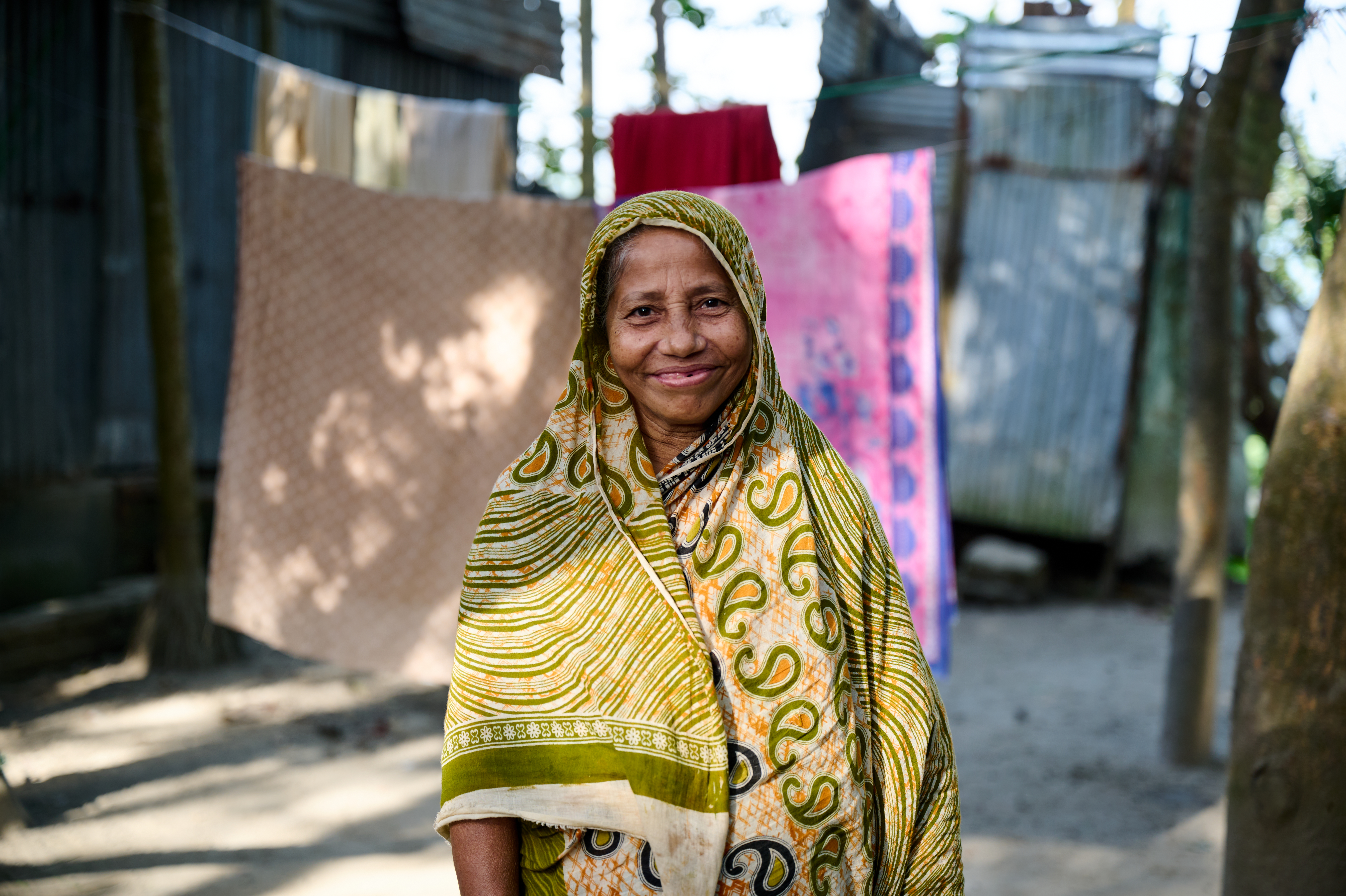 A grandmother smiles to the camera, as washing swaying in the breeze is hung up on a clothes line behind her.